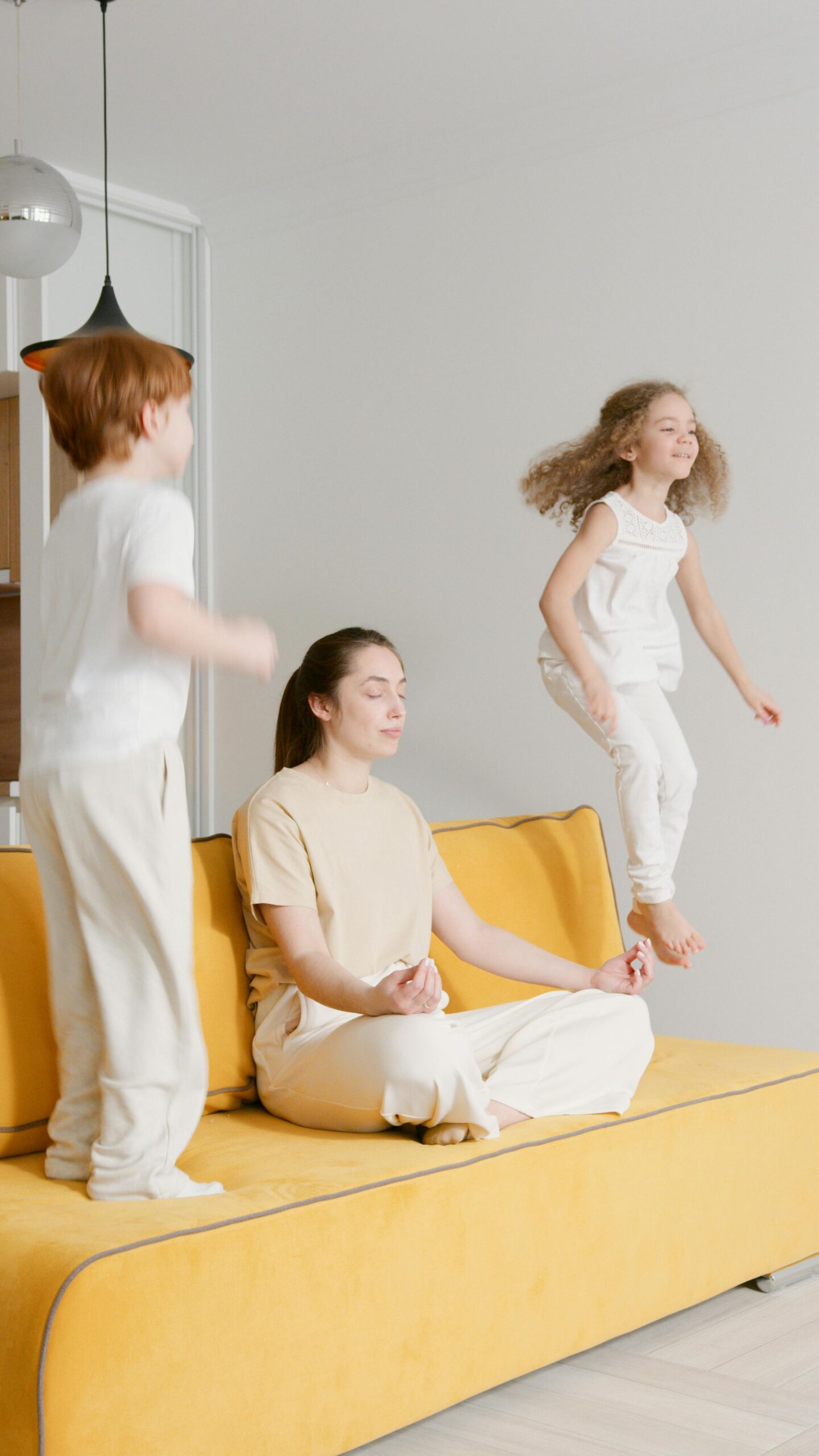 Mother sitting calmly and meditating on a couch while her children jump and play around her, illustrating getting triggered by your child and learning nervous system regulation through conscious parenting.