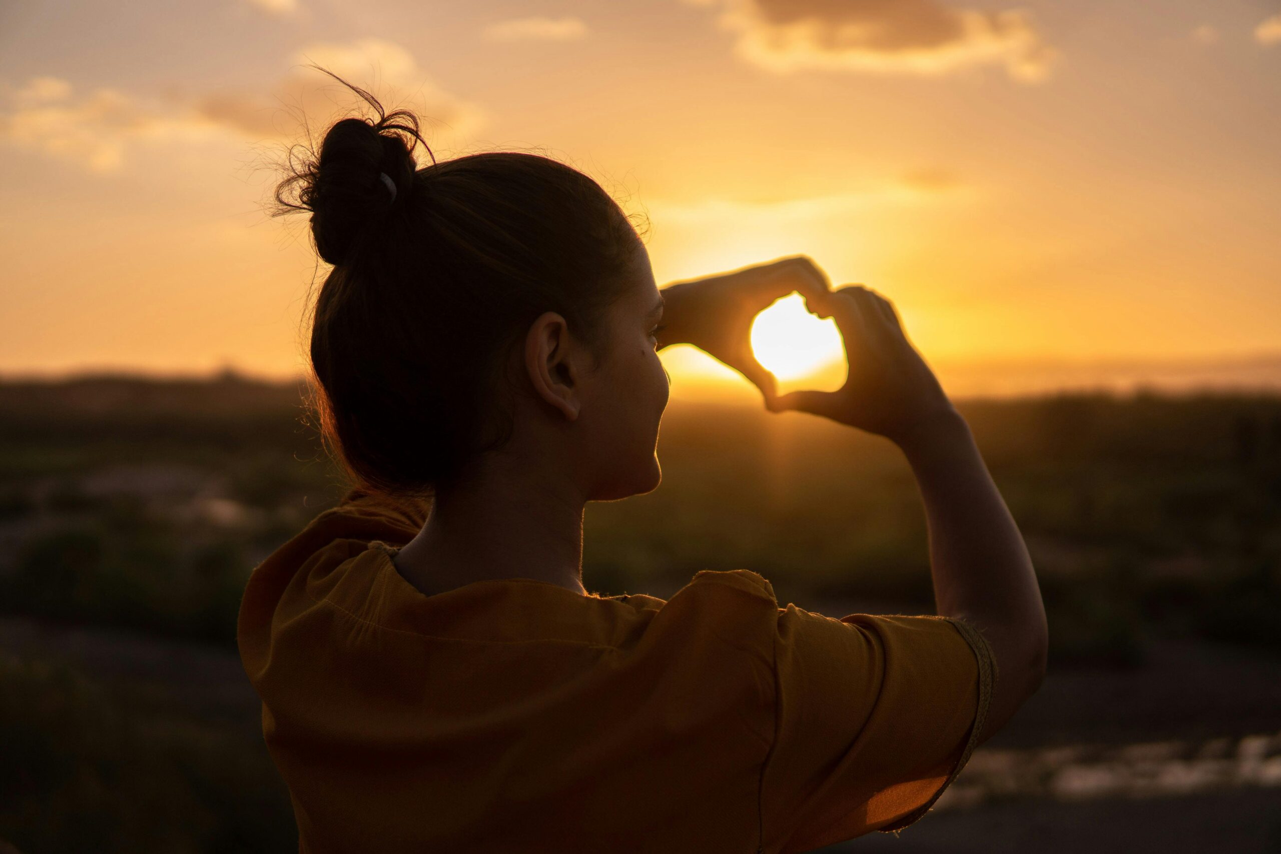 A woman standing outdoors at sunset with her hands raised and forming a heart shape, symbolizing love, hope, and connection.